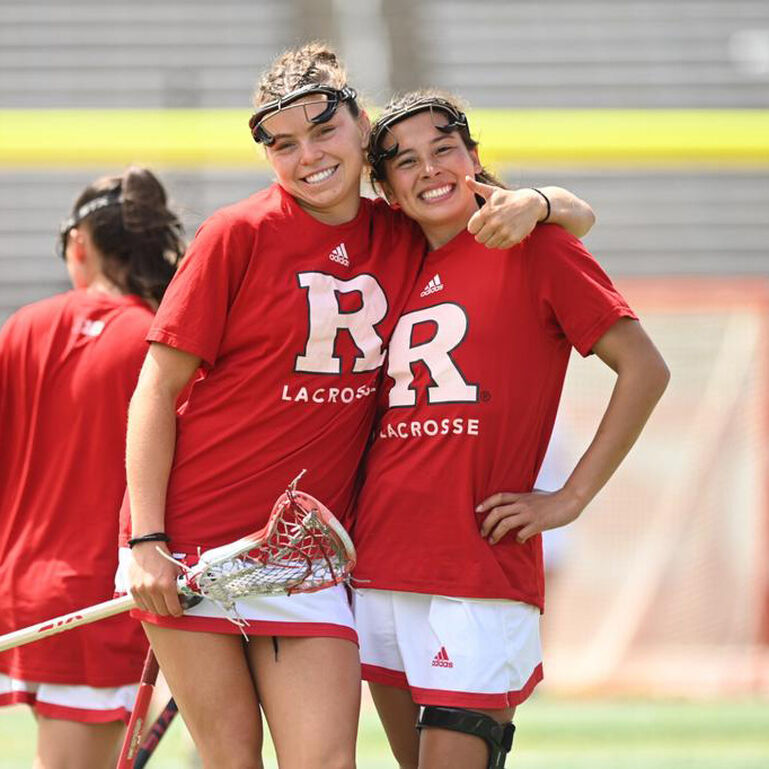 Two Rutgers Women's Lacrosse players smiling at the camera on the field