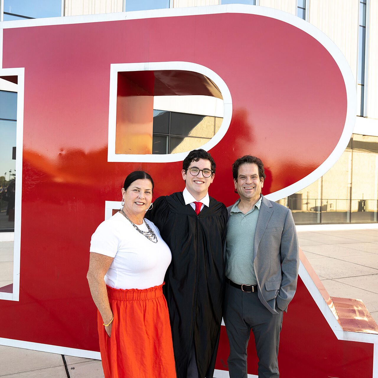 Student in graduation robe standing in front of a large Block R statue with their parents image number 0