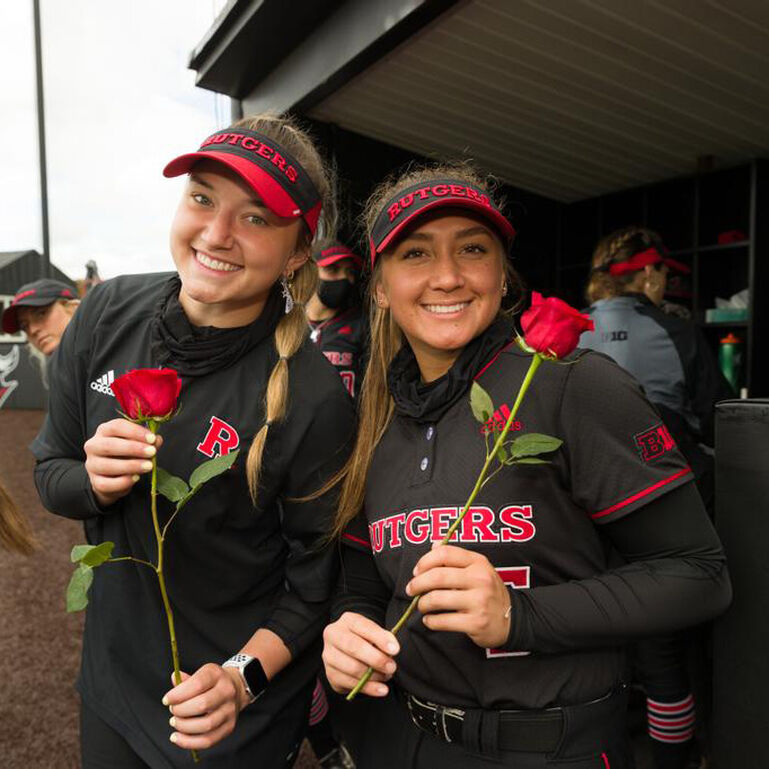 Two Rutgers Softball players smiling and holding roses in front of the dugout